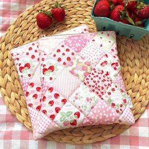 Peut inclure: Scène de pique-nique avec une couverture à carreaux rose et blanc à bords à volants. Un dessous de plat en osier accueille une pochette matelassée à motifs de fraises et un petit panier de fraises fraîches.