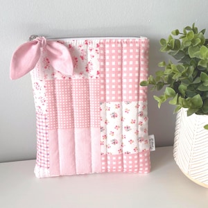 Peut inclure: Une pochette rose matelassée avec un motif patchwork. La pochette présente divers motifs, notamment du vichy, des fleurs et des rayures roses unies. Un nœud rose assorti est attaché à la tirette de la fermeture éclair. La pochette est probablement utilisée pour ranger de petits objets.