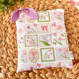 May include: A square fabric pouch featuring a repeating pattern of postage stamp-style illustrations with pink tulips, bows, and hearts on a white background. A lavender fabric bow is attached to the top corner, and a small white tag reads All About Bags co.