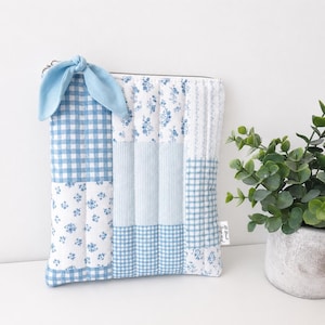 Peut inclure: Une pochette matelassée rectangulaire présentant un motif patchwork de tissus bleu clair et blancs. Les motifs incluent des carreaux vichy, des rayures verticales et de petites fleurs. Un lien en tissu bleu clair est attaché à la fermeture.