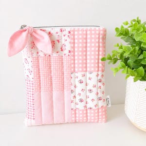 May include: A rectangular quilted pouch featuring a patchwork design of pink and white fabrics in gingham, striped, and floral patterns. A pink fabric bow is attached to the silver zipper pull. A small white tag reads All About Bows.