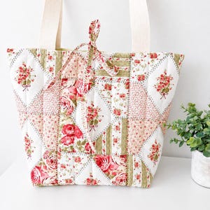Peut inclure: Un sac fourre-tout matelassé présentant un motif patchwork de tissu blanc avec des imprimés de roses roses et des accents rayés vert olive. Le sac comprend deux larges anses en tissu crème et un lien de fermeture en tissu assorti au centre.
