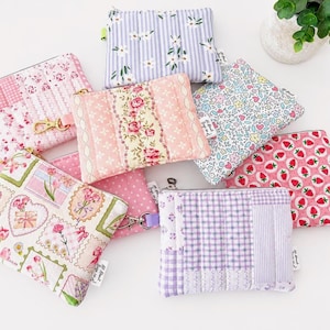 May include: A collection of seven rectangular fabric zipper pouches featuring various patterns including pink floral prints, lavender stripes with white flowers, small colorful blossoms, red strawberries, and purple gingham patchwork designs.