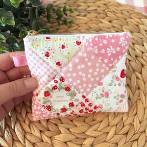 May include: A small, patchwork coin purse with a gold zipper and pink pull tab. The fabric features a variety of pink and white patterns, including polka dots, floral prints, and strawberry motifs. The words "Sweet Strawberry" are printed on the fabric.