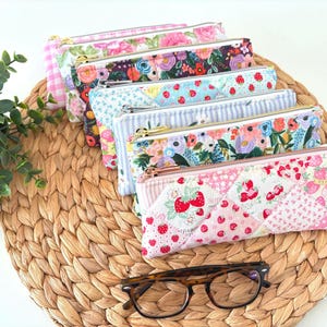 May include: A collection of colorful, patterned pencil cases with gold zippers. Designs include floral, strawberry, and gingham patterns. The cases are arranged on a woven mat, with a pair of tortoiseshell glasses in the foreground.