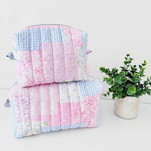 Peut inclure: Deux trousses matelassées rectangulaires empilées verticalement, présentant un motif patchwork de tissus rose clair et bleu. Les motifs incluent des imprimés floraux, des rayures et des carreaux vichy. Chaque trousse a une fermeture éclair.