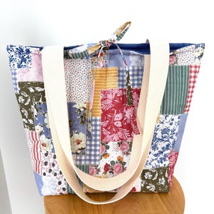 Peut inclure: Un sac fourre-tout matelassé avec un motif patchwork composé de divers carrés de tissu à fleurs, à carreaux vichy et à rayures dans des tons de rose, bleu, vert, jaune et rouge. Le sac comprend deux larges anses en tissu crème et un lien de serrage.