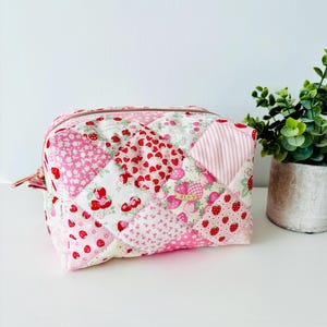 May include: A quilted cosmetic bag with a patchwork design featuring strawberries and hearts in shades of pink and red. The bag has a gold zipper and a small loop. The bag is sitting on a white surface.