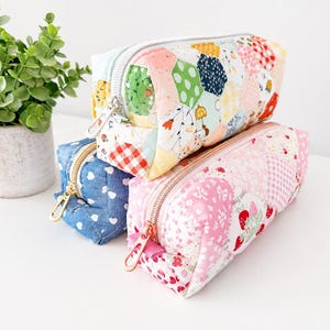 May include: Three rectangular zipped pouches made of quilted fabric. One pouch features a multicoloured hexagonal patchwork pattern, another is solid blue with white hearts, and the third is pink with strawberry and floral patterns.