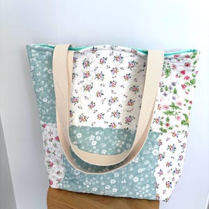 May include: A quilted tote bag featuring a patchwork design of various floral patterns in shades of sage green, white, and pink. The bag has two wide, beige fabric shoulder straps and a solid mint green interior lining visible at the top opening.