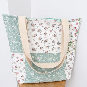 Peut inclure: Un sac fourre-tout matelassé présentant un motif patchwork de panneaux en tissu vert sauge et blanc avec divers imprimés floraux. Le sac comprend deux larges bandoulières en tissu tissé beige et une fermeture éclair vert clair sur le dessus.