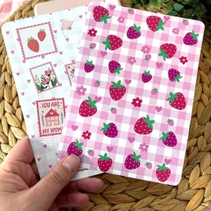 Peut inclure: Deux cartes décoratives avec des motifs de fraises. L'une présente un fond blanc avec des illustrations de timbres-poste de fraises et le texte "YOU ARE MY HOME". L'autre carte a un motif vichy rose avec des graphiques de fraises et de fleurs.
