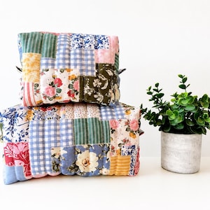 Peut inclure: Deux pochettes rectangulaires matelassées empilées verticalement, présentant un motif patchwork composé de divers tissus à fleurs, à carreaux vichy et à rayures dans des tons bleu, vert, rose, jaune et blanc, avec des coutures visibles sur toute la surface.