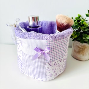 May include: A lavender patchwork fabric cosmetic organiser with a matching bow. It contains a purple perfume bottle, a makeup brush, and a clear clip. A small plant is in the background. The organiser is made of fabric.