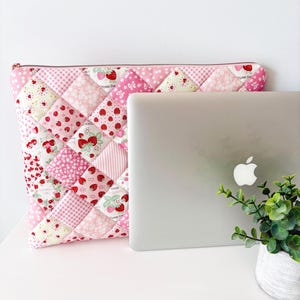 May include: A quilted laptop sleeve featuring a patchwork design of pink and white fabrics with strawberry, floral, and gingham patterns. The rectangular sleeve has a top zipper closure and is positioned behind a silver laptop computer.