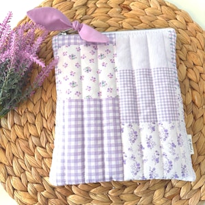May include: A quilted pouch with a lavender bow zipper pull. The pouch has a patchwork design with floral, gingham, and striped patterns in white and lavender. The pouch is on a woven, circular placemat.