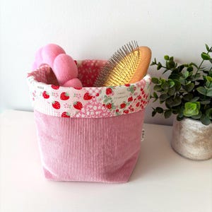 May include: A pink corduroy storage basket with a strawberry print lining. The basket contains a pink hair accessory and a wooden-handled hairbrush. The rim of the basket has a strawberry and floral pattern. The text "Sweet Strawberry" is visible.