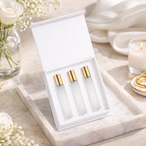 May include: A white gift box with an open lid, revealing three frosted glass perfume bottles with gold-colored caps. The box sits on a marble tray, with a candle and flowers in the background.