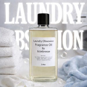 Könnte beinhalten: Eine transparente Glasflasche mit Laundry Obsession Duftöl von Eclatbreeze, mit schwarzem Verschluss. Das Etikett listet Duftnoten und das Volumen als 100 ml auf. Der Hintergrund umfasst weiße Handtücher und ein blaues Tuch.