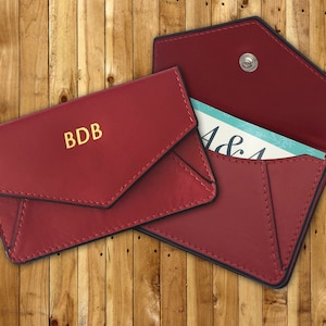 May include: Two red leather envelope style card holders. The top holder has the initials "BDB" embossed in gold. The bottom holder is open and shows a blue and white card inside.