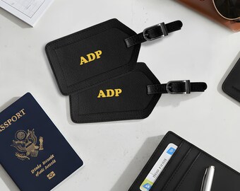 May include: Two black textured luggage tags with the letters ADP embossed in yellow. Each tag features a black strap with a silver-toned metal buckle. The tags are positioned near a dark blue passport, a black card holder, and sunglasses.