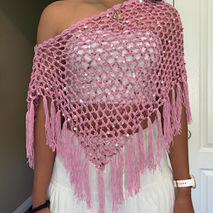 May include: A pink crochet poncho featuring an open net pattern with integrated shimmering sequins and long fringe trim along the bottom edge. The garment is designed with an asymmetrical neckline that rests over one shoulder, creating a triangular shape.