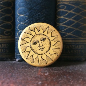 May include: A round, yellow button with a smiling sun design. The sun has a face with eyes, a nose, and a mouth. The button is set against a backdrop of dark blue books with gold lettering.