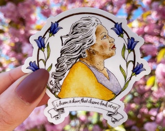 May include: A white sticker featuring a watercolour illustration of a woman with grey hair and a yellow shirt. The woman is surrounded by blue flowers and green leaves. The text "I dream a dream that draws back to me" is written on a white banner at the bottom of the sticker.