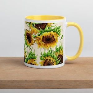 Puede incluir: Una taza de cer&aacute;mica blanca con un borde y un asa amarillos. La taza presenta un patr&oacute;n repetitivo de girasoles amarillos con hojas verdes sobre un fondo crema.