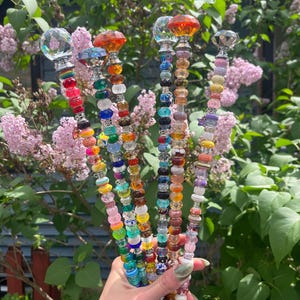 Peut inclure: Une collection de piquets de jardin perlés colorés avec des dessus décoratifs en cristal et en forme de gemme. Les piquets présentent une variété de perles dans les tons de rouge, orange, jaune, vert, bleu et violet. Le fond comprend du feuillage vert et des fleurs roses.
