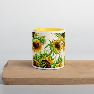 Puede incluir: Una taza de cer&aacute;mica blanca con un borde amarillo presenta un patr&oacute;n repetitivo de girasoles con hojas verdes sobre un fondo crema.