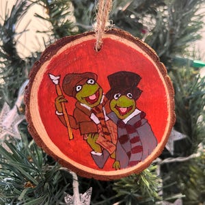 Puede incluir: Adorno navideño de madera pintado a mano con dos personajes de rana verde con ropa de invierno. El adorno tiene un fondo rojo y un borde de madera natural. Se adjunta una cuerda de yute para colgarlo.