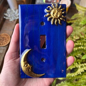 May include: A decorative blue light switch cover featuring a gold sun and crescent moon design. The switch plate has a glossy finish and a rectangular opening for the switch. The sun and moon are raised, adding a 3D effect.