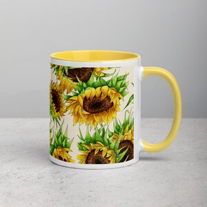 Puede incluir: Una taza de cer&aacute;mica blanca con un borde y un asa amarillos. La taza presenta un patr&oacute;n de girasoles amarillos con hojas verdes sobre un fondo crema.