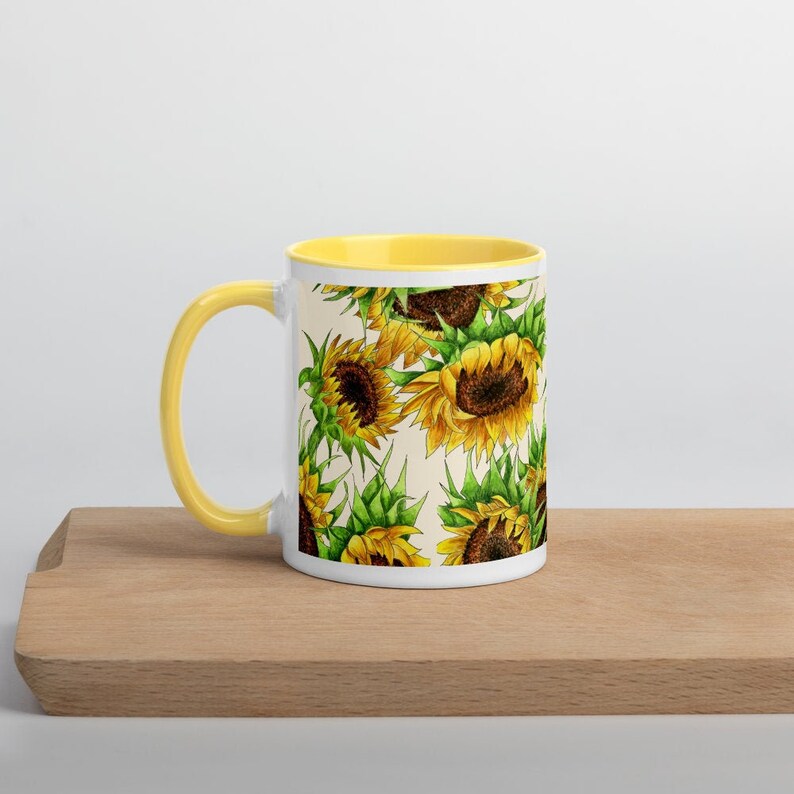 Puede incluir: Taza de cer&aacute;mica blanca con borde amarillo y un patr&oacute;n de girasoles sobre un fondo crema.