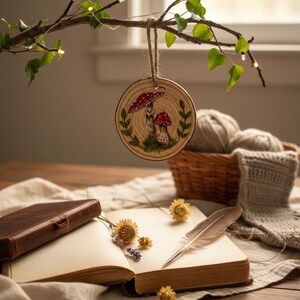 May include: A wooden ornament with a mushroom design hangs from a branch with ivy and fairy lights. An open book, dried flowers, and a feather quill rest on a linen cloth. A basket with yarn and a knitted item are in the background.