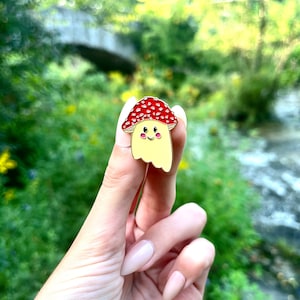 May include: A red and white enamel pin of a smiling mushroom with a yellow body and a red and white cap. The pin is being held by a hand in front of a blurred background of green foliage.
