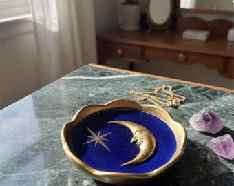 May include: A small, round, scalloped-edge trinket dish with a deep blue interior featuring a raised gold-toned crescent moon with a face and a gold-toned four-pointed star. The dish has a metallic gold rim and a smooth, glossy finish.