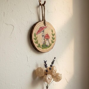 May include: A round wooden wall hanging featuring a hand-painted mushroom design. The mushrooms have red and white caps, surrounded by green leaves. The wood slice is suspended by a brown rope. A small bouquet of dried flowers is below.