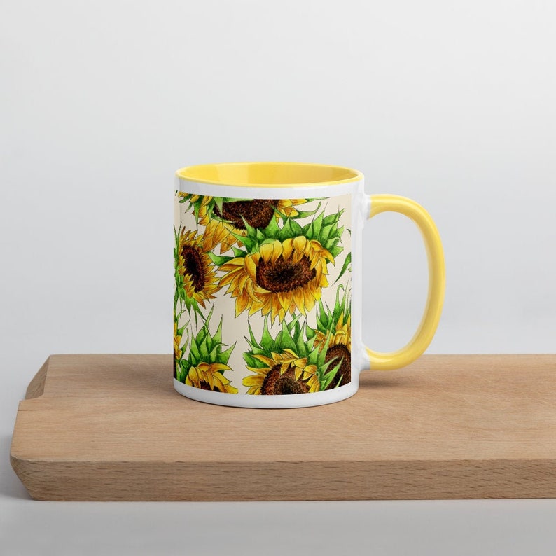 Puede incluir: Una taza de cer&aacute;mica blanca con un borde y un asa amarillos. La taza presenta un patr&oacute;n repetitivo de girasoles amarillos con hojas verdes sobre un fondo crema.
