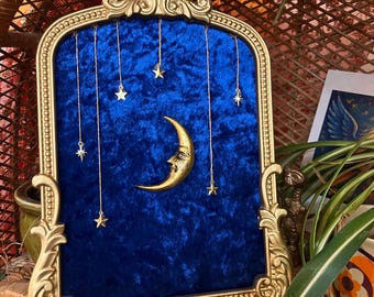 Puede incluir: Un marco decorativo arqueado con un acabado dorado ornamentado que contiene un fondo de terciopelo azul intenso. Una luna creciente dorada con rostro está centrada, rodeada por seis pequeños dijes de estrellas doradas en cadenas finas.