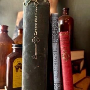 May include: A vintage-style key bookmark hangs from a dark green book. The key is gold-colored and attached to a chain. Other books and brown bottles are in the background, creating a classic, antique aesthetic.