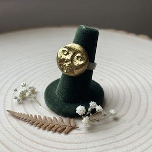 May include: A gold-colored ring with a textured face design, displayed on a dark green ring holder. The ring is set against a light wood background, with dried flowers and a fern leaf.