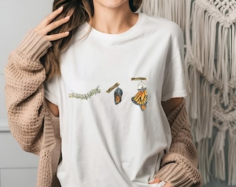 May include: White t-shirt with a graphic of a caterpillar, chrysalis, and butterfly. The graphic is a watercolor style illustration.