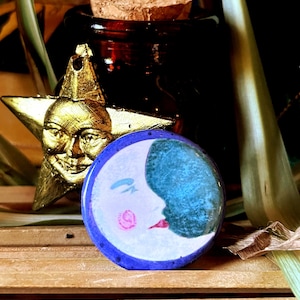 May include: A circular pin featuring a stylized moon face with a pale white complexion, a blue crescent shadow, a pink cheek, and red lips, framed by a dark blue border. Beside it sits a gold-colored star ornament with a detailed human face.