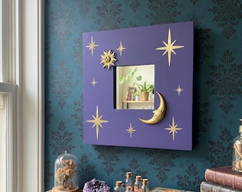 May include: A square, purple wall mirror with a central square mirror. Gold-colored sun, moon, and star embellishments adorn the frame. The mirror is mounted on a teal wall with a dark floral pattern.