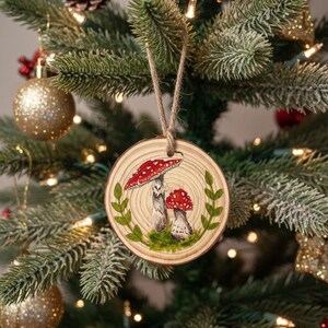May include: A round wooden Christmas ornament featuring a hand-painted design of two red and white mushrooms with green leaves. The ornament is suspended by a natural twine string and hangs on a decorated Christmas tree.