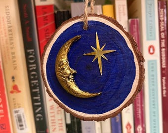 Pode incluir: Um ornamento circular de fatia de madeira com um centro pintado de azul profundo, uma lua crescente dourada tridimensional com um rosto e uma estrela dourada de quatro pontas. O ornamento pende de um laço de corda de juta natural.