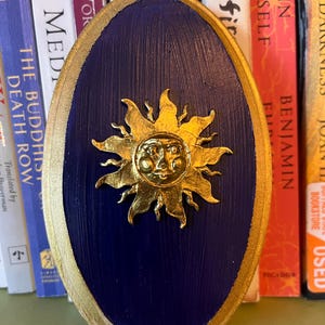 May include: An oval-shaped plaque with a deep blue painted centre and a metallic gold border. Centred on the plaque is a three-dimensional gold sun emblem featuring a human-like face with rays extending outwards in a circular pattern.