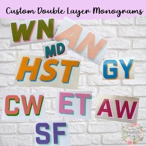 May include: A collection of custom double-layer monograms in various color combinations, including green, orange, pink, and blue. The monograms are displayed against a white brick wall. The text "Custom Double Layer Monograms" is at the top.